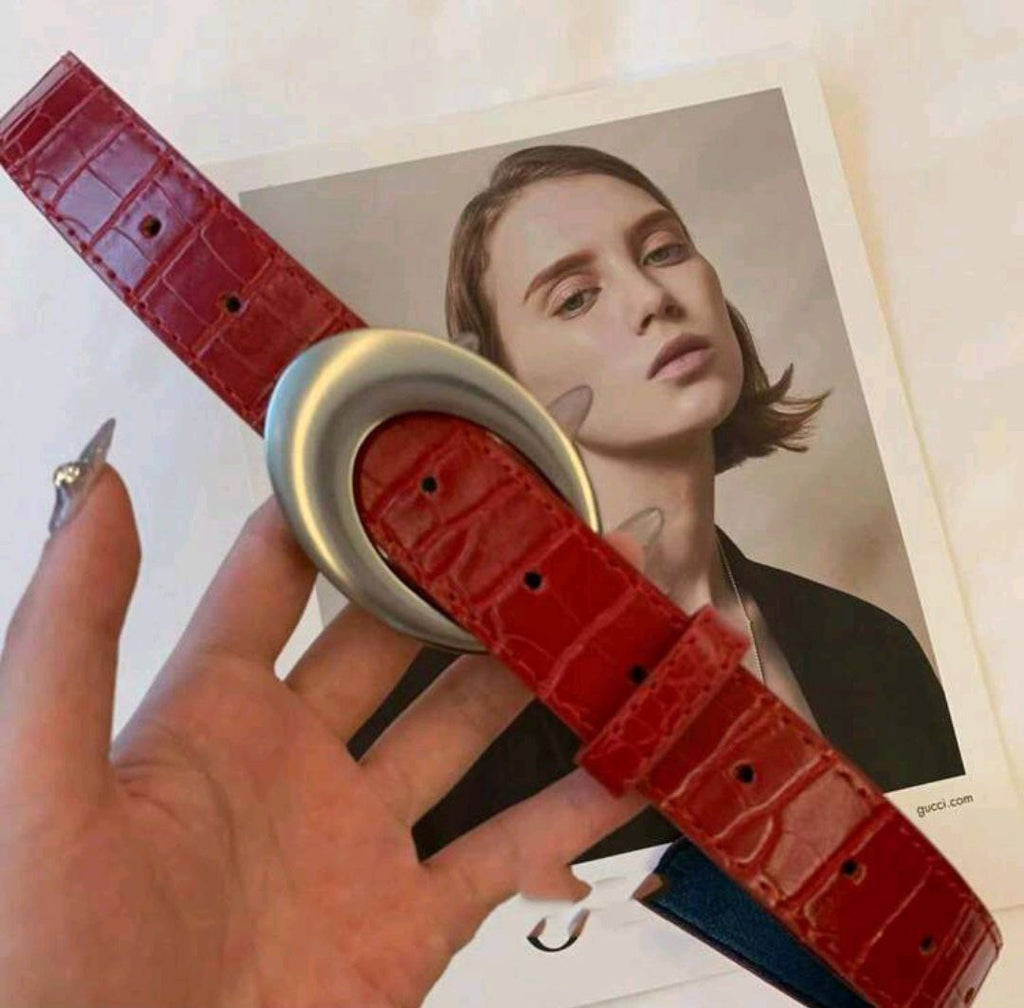 Red belt with a large silver buckle held by a hand, with a blurred background of a magazine cover featuring a person.